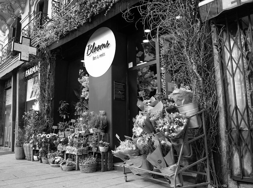 Exterior view of Bloom's flower shop on a city street, showcasing a window display filled with a variety of fresh floral arrangements and bouquets. The display includes wrapped flower bunches featurin