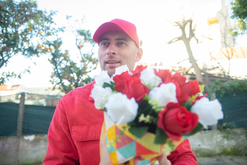 A delivery person dressed in a red uniform and matching cap holding a vibrant bouquet of fresh flowers, featuring red and white roses with green foliage, wrapped in colorful patterned paper. The perso
