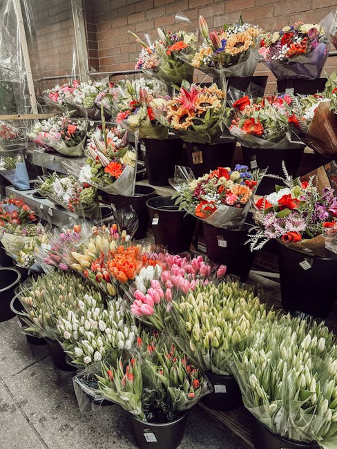 An outdoor display of numerous fresh flower bouquets arranged in black buckets, featuring a variety of spring blooms such as pink and white tulips, orange and red roses, and mixed seasonal flowers wit