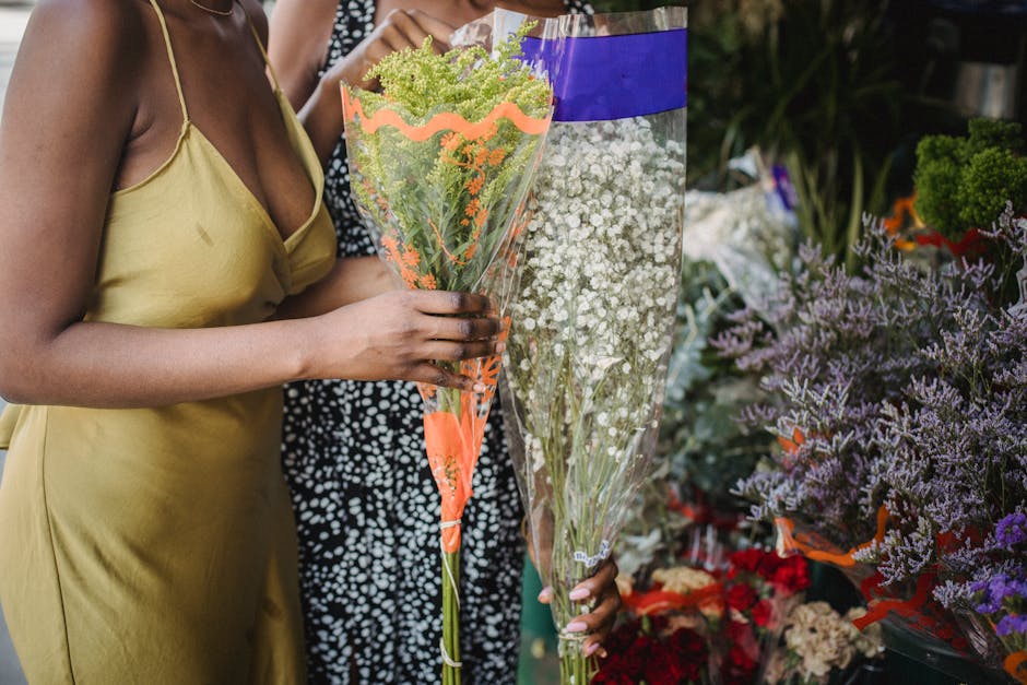A person wearing a yellow satin dress holds a tall bouquet wrapped in orange and transparent floral paper, featuring green and purple flowers with delicate green foliage. The person stands near a flow